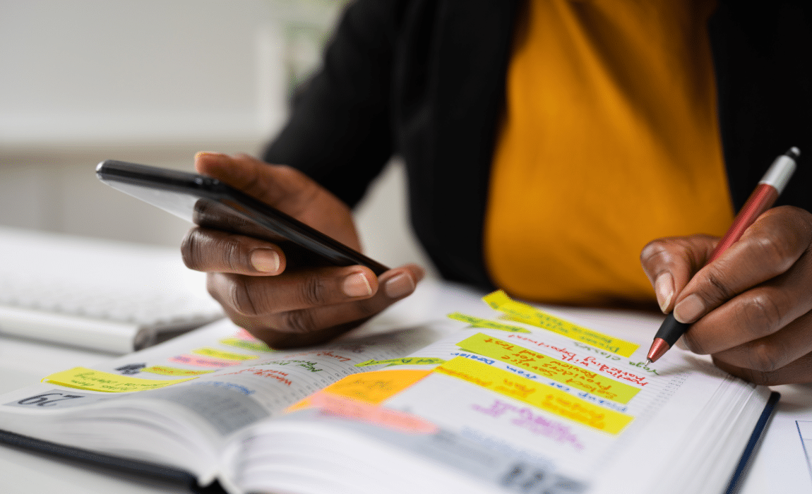 Close up image of someone's hands. One hand is holding a phone and the other is writing something in a calendar