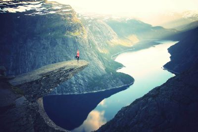 Photo of woman in a yoga tree pose, on top of a rocky ledge, surrounded by mountainous scenery, with a lake or river far below her.
