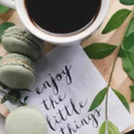 Birds eye image of a cup of coffee on a wooden board, next to some foliage, with green macarons, and a note saying 'enjoy the little things'.