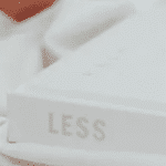 Photo of a white book placed on white sheets or a table cloth with the word 'Less' printed on the book's spine. Close to the book in the background part of a peach-coloured mug can be seen.