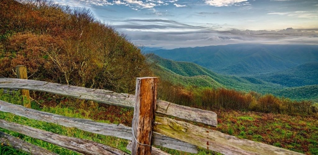 Outdoor scene of rolling green hills, with low cloud in the far distance, and a wooden fence in the foreground.